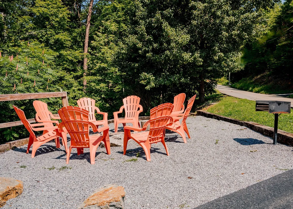 A circular arrangement of vibrant red Adirondack chairs on a gravel surface, surrounded by trees. The setup invites guests to gather around a campfire, socialize, and experience outdoor fun together.