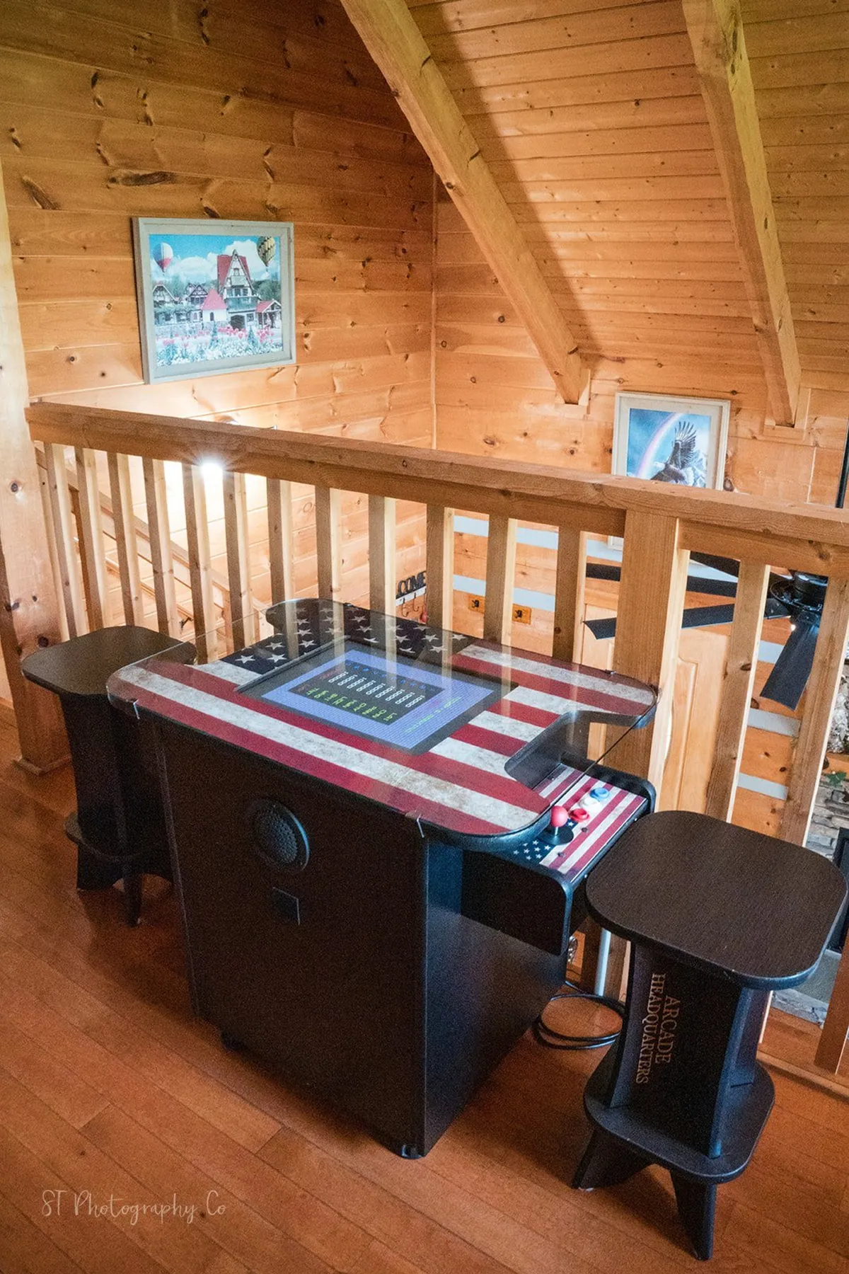 This image captures the open loft game room, featuring an arcade table with an American flag design, a rustic wooden railing, and framed artwork on the wall. Natural light streams in from large windows, brightening the playful scene.
