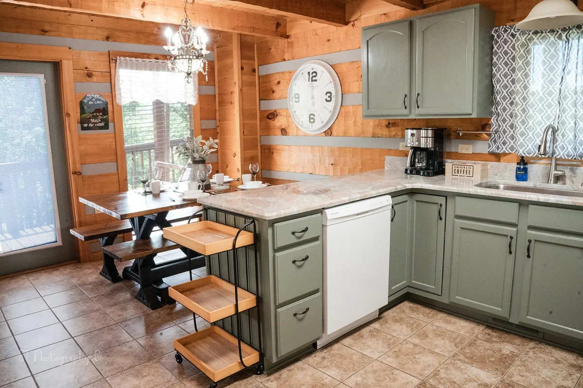 A warm, inviting kitchen and dining space featuring a substantial wooden dining table, a kitchen island, elegant chandelier, and a large clock. The table is set to accommodate a large group, and the kitchen is well-equipped for preparing meals.