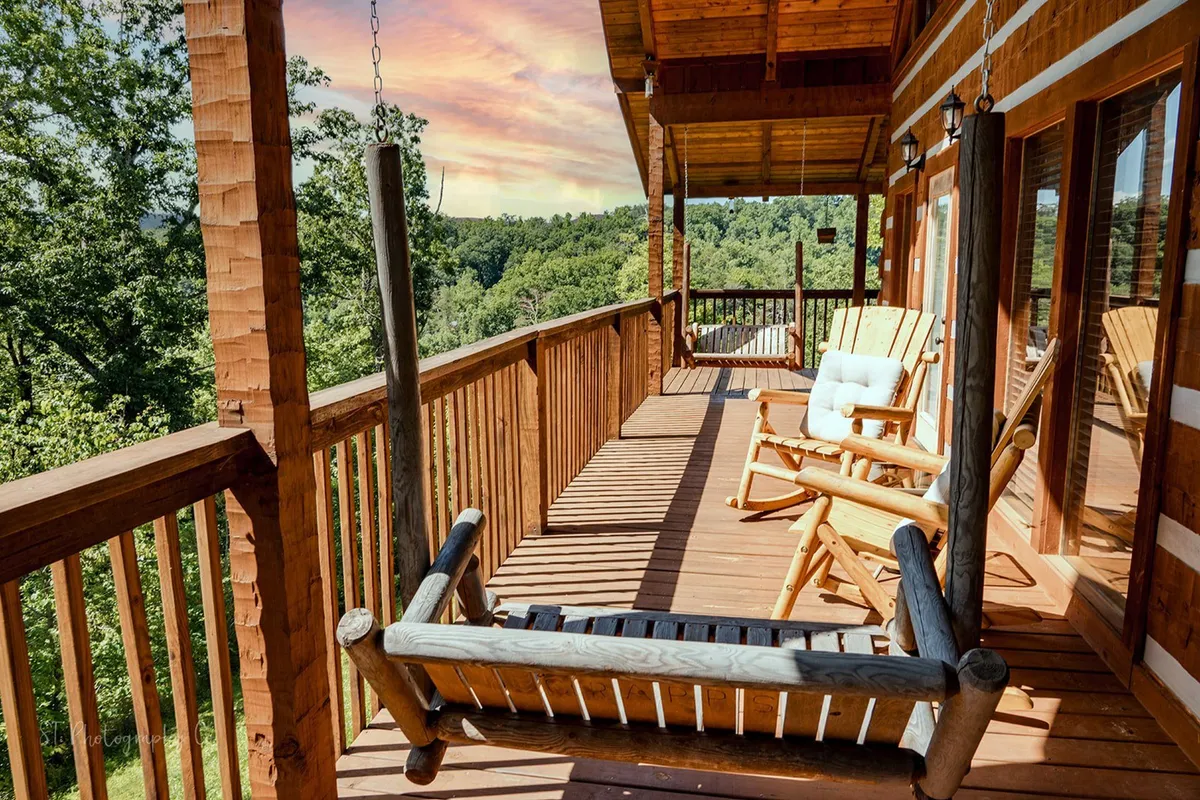 A scenic portion of the wrap-around porch featuring a swing bench, several wooden rocking chairs, and a sturdy wooden railing. The seating is oriented to provide perfect views of the surrounding trees and sunsets. The area looks serene and is ideal for relaxing with a book or conversing while soaking in the sounds of nature.