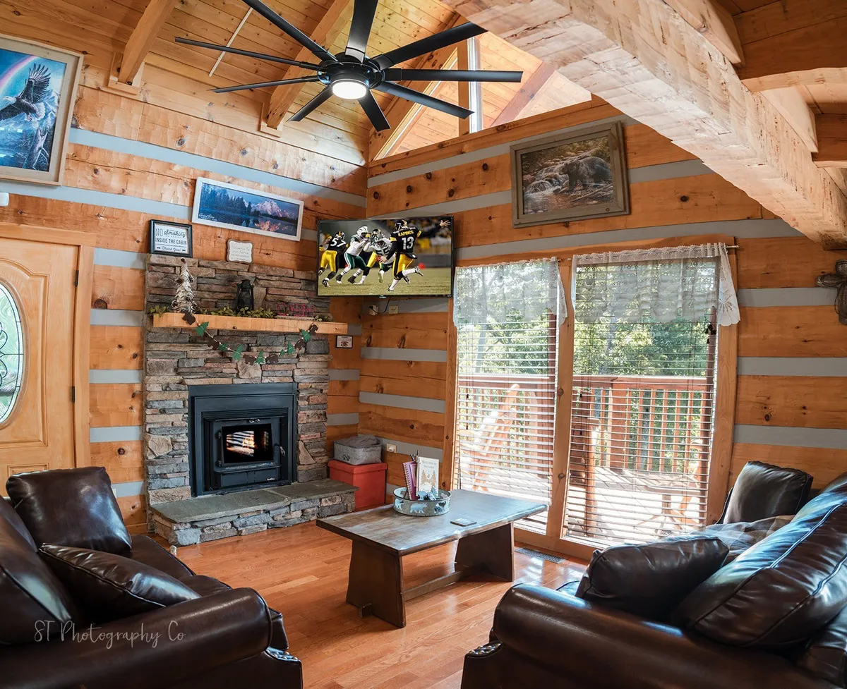 A bright, open living area centered around a crackling stone fireplace with plush leather sofas arranged for conversation. Large windows let in plenty of natural light, and a flat-screen TV is visible above the mantle, making this a perfect spot for movie nights or cozying up by the fire.