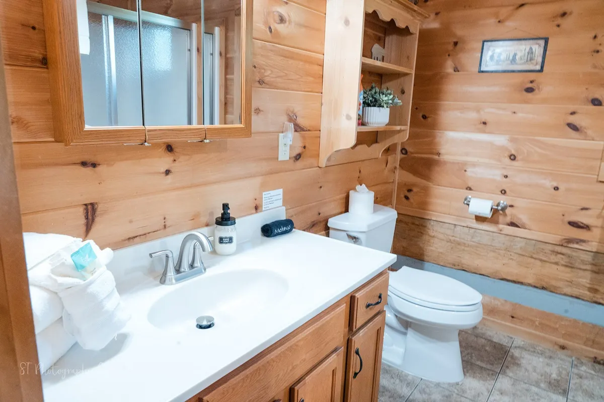 This image shows a comfortable bathroom with a vanity, sink, mirror, wooden cabinetry, shelving with plants, toilet, and warm natural light. The spacious design and practical layout emphasize ease of use for guests with varying mobility needs.