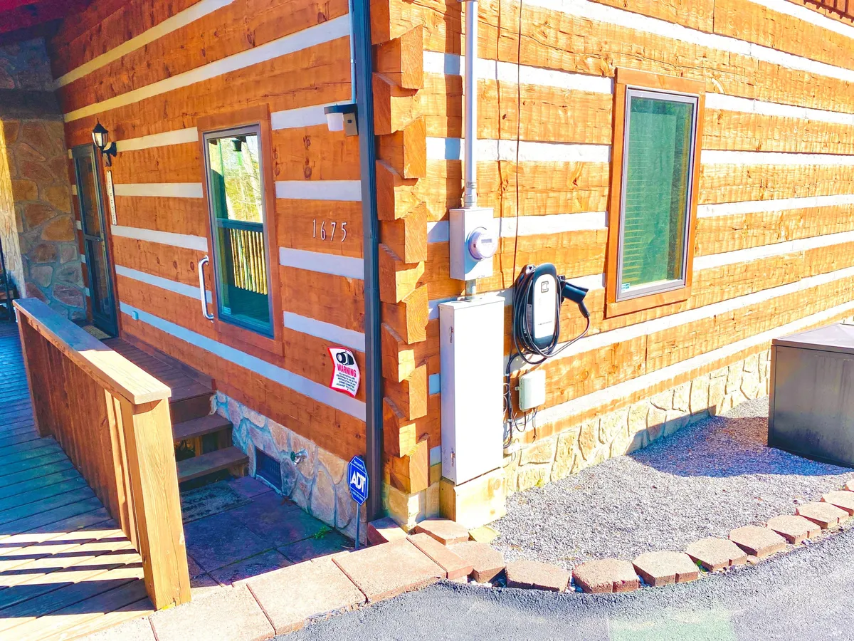 A welcoming view of the log cabin's exterior, showing its front steps, inviting porch, and windows surrounded by natural greenery. The setting conveys a tranquil retreat nestled in nature, perfect for enjoying the outdoors.