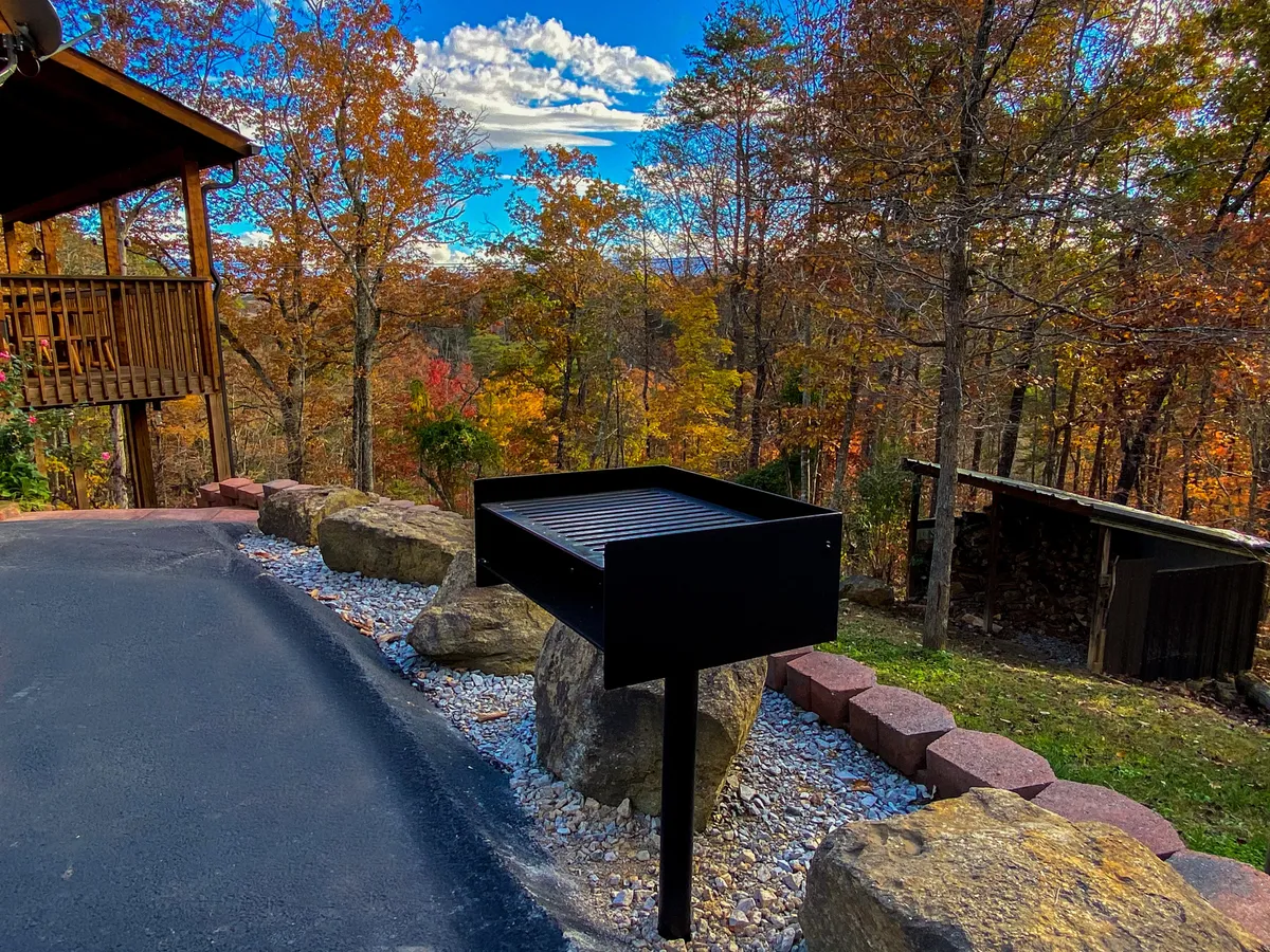 An outdoor grilling area situated on a deck, surrounded by tall trees and accessed via a stone pathway. The grill sits ready for use, with ample space for group barbecues, outdoor meals, or simply enjoying the fresh air. The deck area is integrated into the natural landscape, perfect for al fresco dining or sunset gatherings.