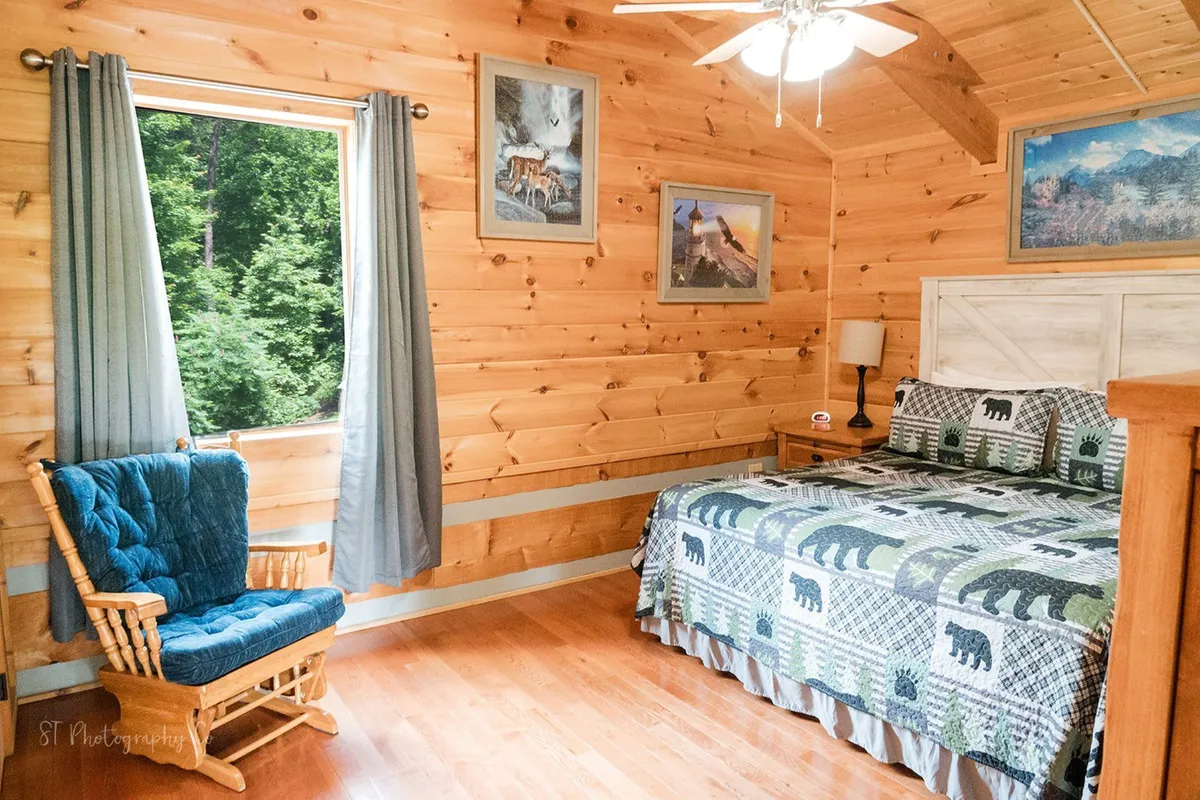 A tranquil bedroom with bear-themed bedding, a wooden rocking chair, nature-inspired artwork, and warm wood-paneled walls. Sunlight streams in from a window, creating a peaceful and inviting retreat ideal for restful nights or quiet mornings.