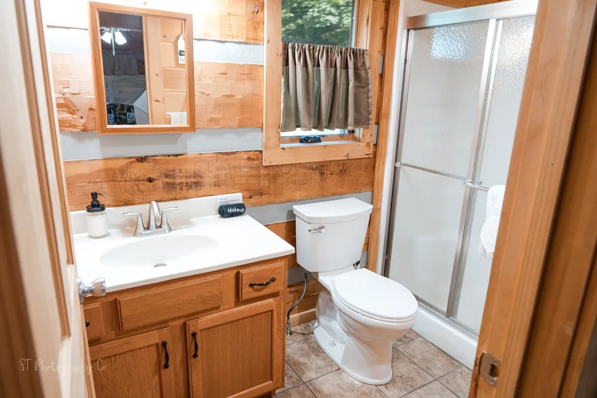 A warmly lit private bathroom featuring wooden paneling, a sink area, a walk-in shower, and a mirror. The modern yet rustic ambiance invites relaxation after a day of adventure, with all linens provided for a spa-like experience.