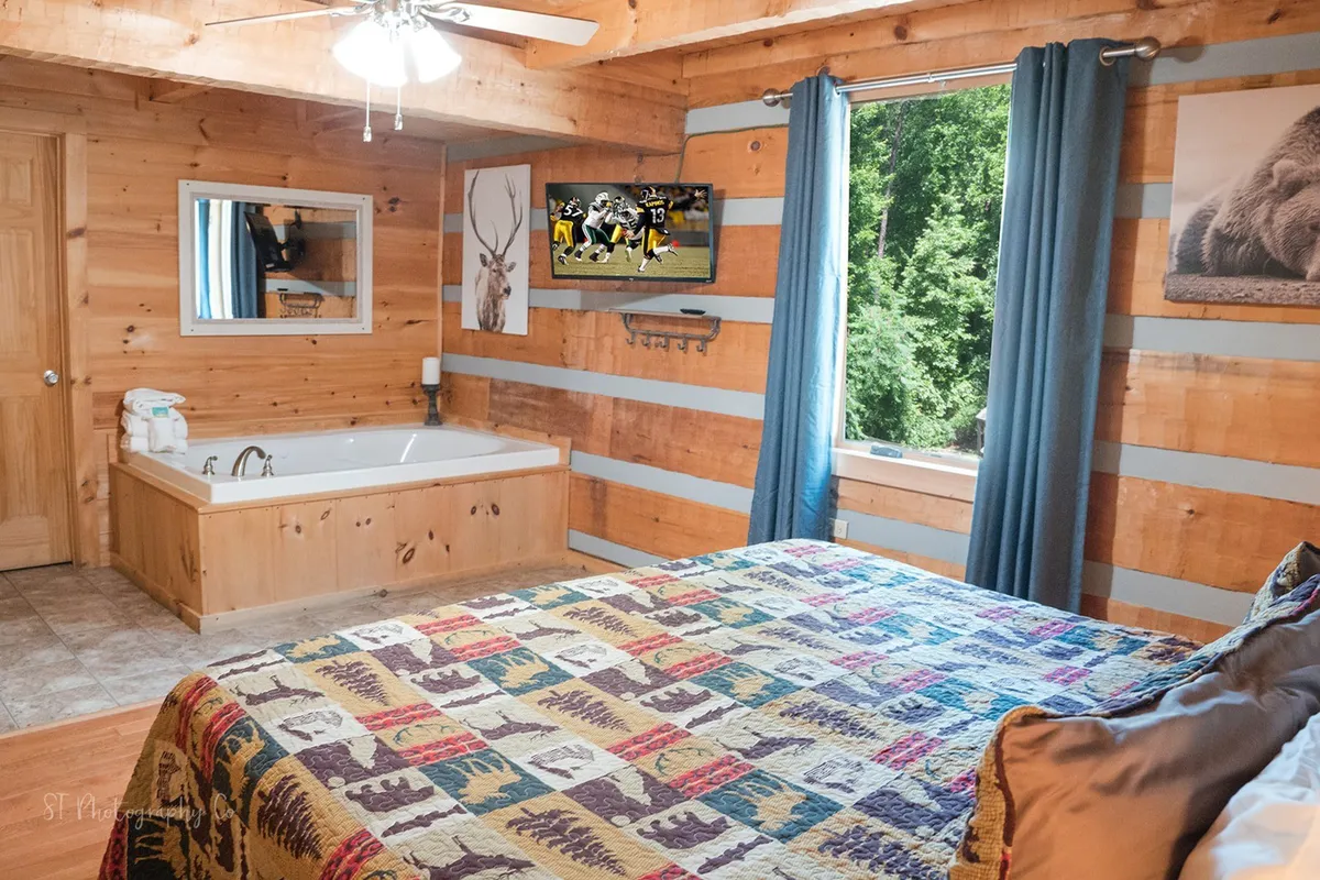 A cozy bedroom featuring a bed with a colorful quilt, a whirlpool (jacuzzi) tub, a wall-mounted television, and a large window with views of the outdoors. The space is bright and inviting, with plenty of natural light and rustic accents, perfect for relaxation or catching up on favorite shows.
