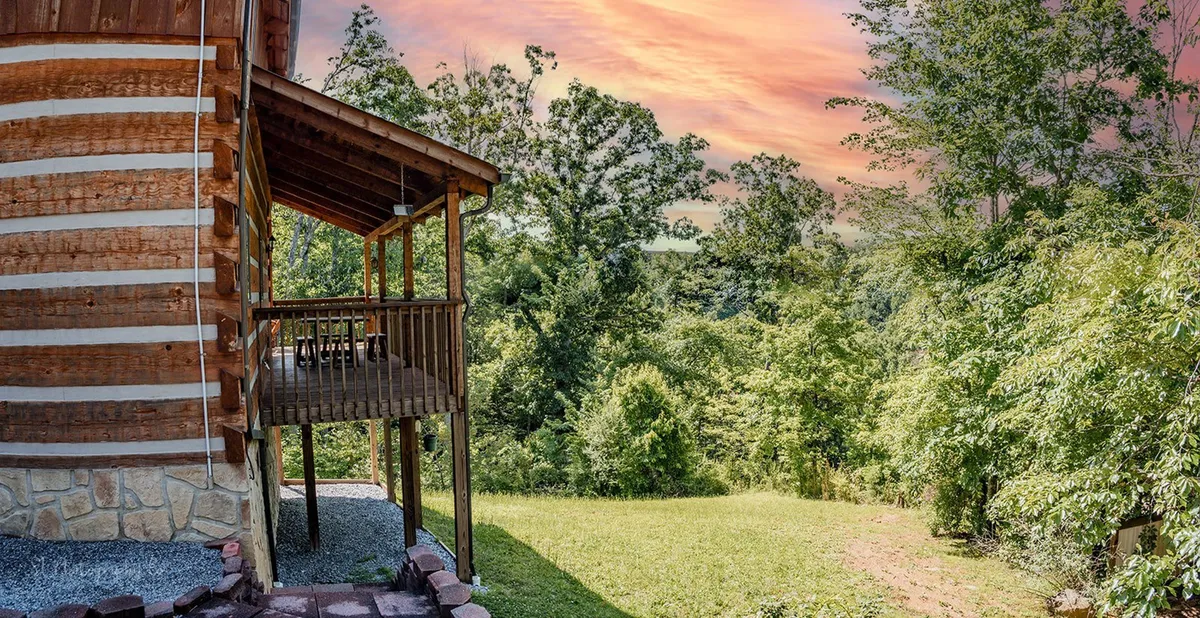 A classic log cabin structure set amid lush greenery, with a spacious deck area overlooking distant hills. The scene captures a peaceful outdoor environment perfect for family gatherings, relaxation, and enjoying nature.