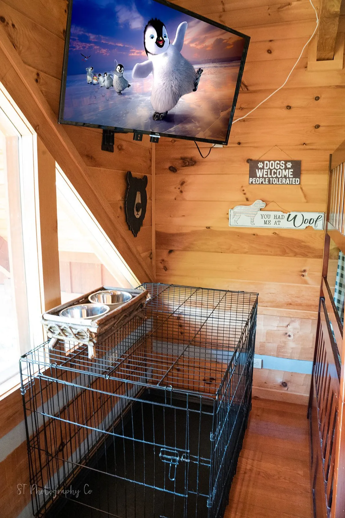 A cozy corner inside the cabin with a television displaying animated penguins, a prominent dog crate, and dog food bowls. Decorative wall signs add a playful, homey touch.