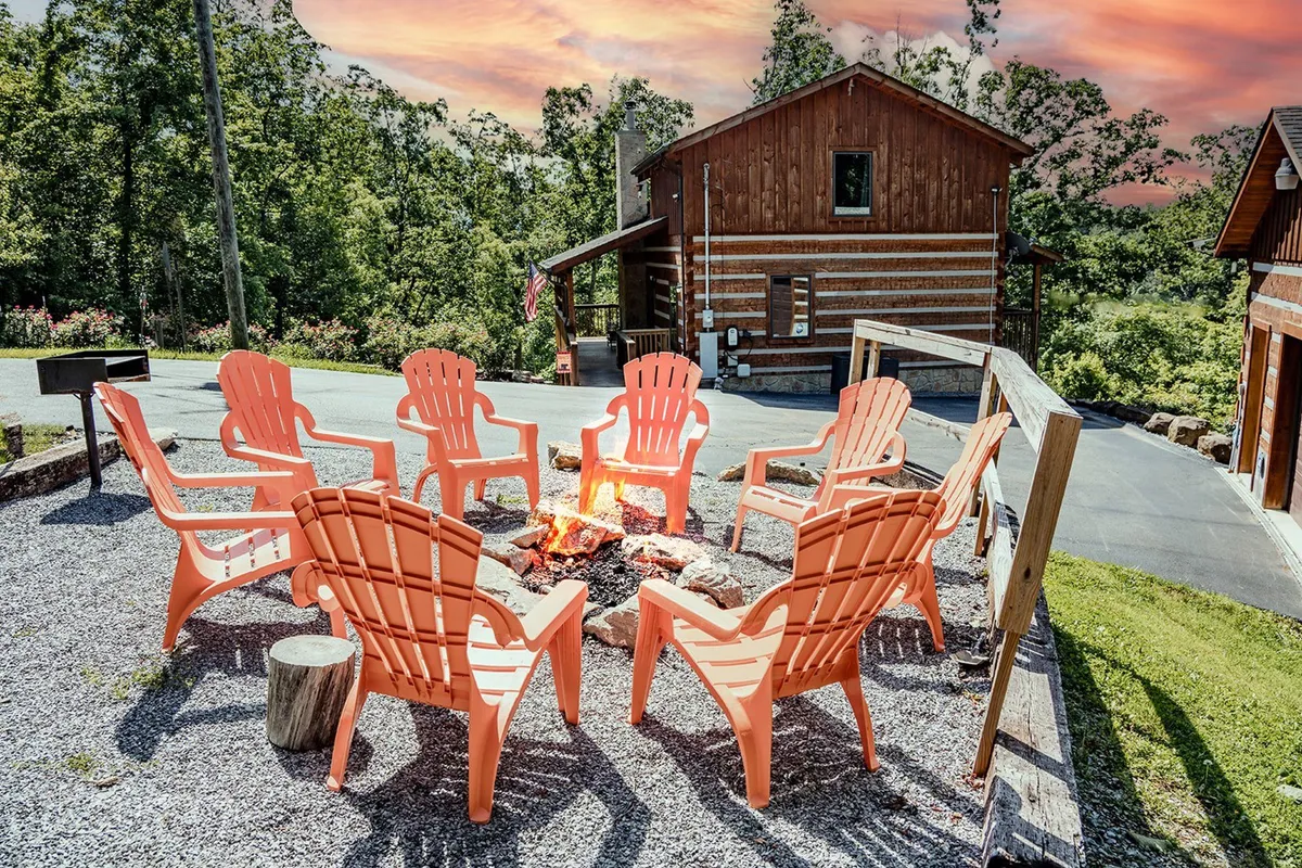 A cozy fire pit area just outside the cabin, with a circle of orange chairs and rustic log seating arranged for group gatherings. The cabin structure is visible in the background, providing a sense of location and context. The area feels inviting—ready for marshmallow roasting, storytelling, and connecting under the stars.