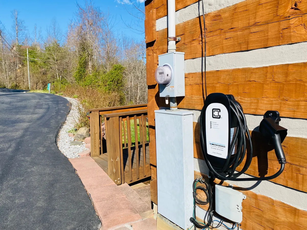 A close-up exterior shot featuring the electric vehicle charger mounted on the wooden deck, with natural landscaping in the background. The image highlights the convenience of EV charging and the eco-friendly amenities, while still keeping the outdoor, natural feel of the property intact.
