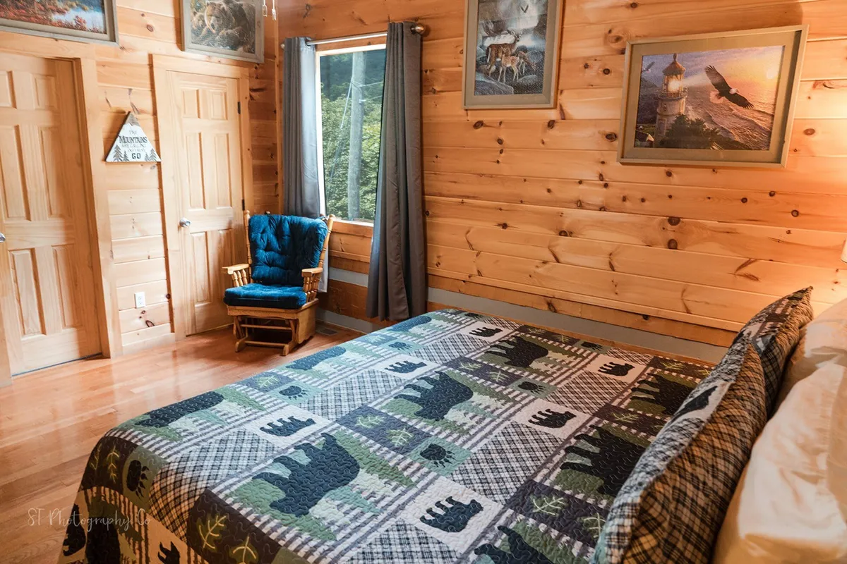 A sunlit nook featuring a bed with a bear-patterned quilt and a blue rocking chair placed near a window, letting in natural light and providing a view of the outdoors. The setup evokes a cozy, inviting ambiance ideal for sipping coffee while enjoying the scenery.