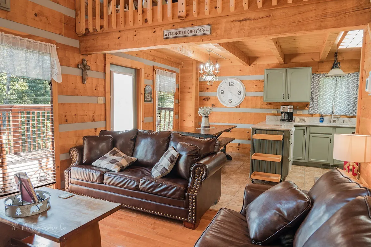 A wide-angle view that brings together the living area, kitchen, and dining space. The image shows leather sofas, wooden beams, a chandelier above the dining area, and the kitchen in the background, illustrating the open-concept layout.