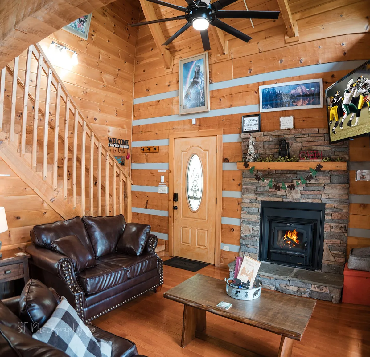 A warm and welcoming living room with leather sofas gathered near a stone fireplace, a TV screen, a wooden staircase, and the front door in view. The space is set up for relaxation, gathering, or catching up on entertainment, with room for additional guests.