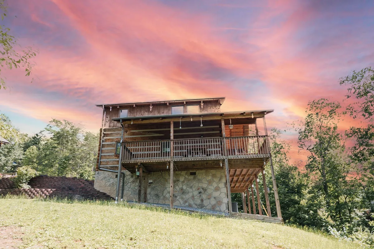 A wide shot of the cabin's wooden deck set atop a stone foundation, bathed in the warm glow of a vibrant sunset sky. The setting offers plenty of space for gatherings and relaxation, with panoramic views of the surrounding mountains and sky.